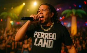 A man with dreadlocks and a gold chain raps into a microphone at a concert with inflatable palm trees in the background.