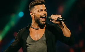 Ricky Martin sings passionately into a microphone on stage, wearing a black shirt and chain necklace.