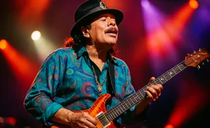 Carlos Santana plays his guitar on stage, wearing a hat and a colorful shirt.