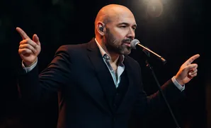 A bald man in a suit sings into a microphone on a dark stage, with his hands gesturing.