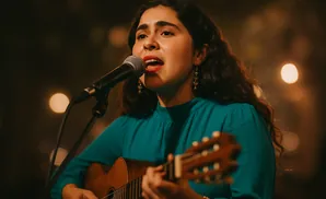 A woman with dark, curly hair sings into a microphone while playing an acoustic guitar.