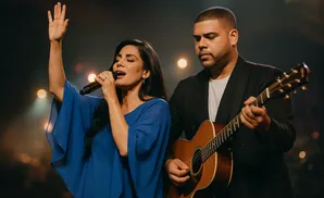 A woman sings into a microphone with her arm raised while a man plays guitar.