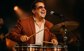 A smiling male drummer in a brown suit plays drums on stage with a microphone and cymbal.