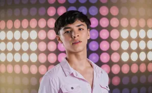 Young man with dark hair and braces, wearing a pink shirt, poses against a background of blurred pink and purple circles of light.