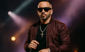 A man wearing sunglasses, a bomber jacket, and a cross necklace stands on stage with spotlights behind him.
