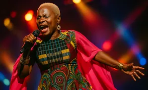 Singer with closed eyes belts into a microphone, wearing a colorful dress and pink cape, bathed in stage lights.