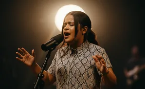 A woman sings passionately into a microphone, eyes closed, with hands raised in a dark, atmospheric setting.