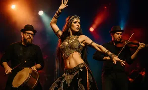 A belly dancer performs with a violinist and drummer on a dimly lit stage with colorful spotlights.