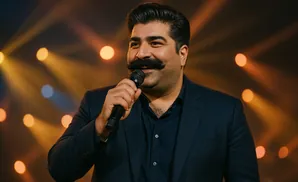 A man with a large mustache, wearing a dark suit and shirt, speaks into a microphone on a brightly lit stage.