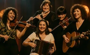 A group of five women playing musical instruments, including violins, a flute, an accordion, and a guitar.