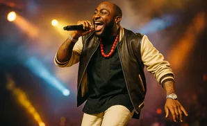 A Black man in a black and tan jacket sings into a microphone on stage with colored stage lights.