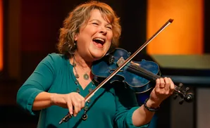 A woman with short, curly hair laughs joyfully as she plays a blue violin.