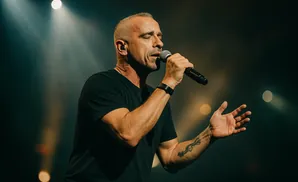 Man singing into a microphone on a dark stage with spotlight and blurred lights.