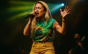 A woman with blonde hair sings into a microphone on stage, wearing a tie-dye shirt and yellow shorts.