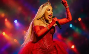 A drag queen in a sparkly red dress and crown sings into a microphone with a fist raised.