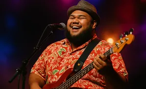 A man in a floral shirt and fedora sings into a microphone while playing a bass guitar on stage.