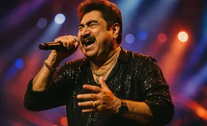 Man singing into a microphone on stage, wearing a sequined shirt and gold jewelry.