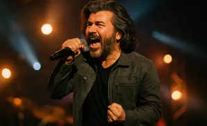 A man with long, dark hair and a beard sings into a microphone on a dimly lit stage.