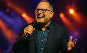 A man in a suit and glasses holds a microphone, speaking with a wide smile and open hands.
