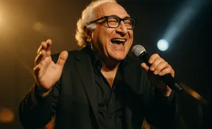 A man with white hair and glasses sings into a microphone, gesturing with his left hand.