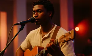 A Black man plays an acoustic guitar and sings into a microphone on a dimly lit stage.