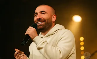 Man with a beard smiles and holds a microphone while wearing a light-colored hoodie.
