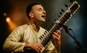 Man in a gold outfit playing a sitar with a microphone in front of him.