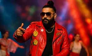 A man with a bun hairstyle and beard wears a red leather jacket with roses and a white star, pointing his finger on stage.