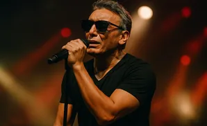 Man in black t-shirt and sunglasses singing into a microphone on stage.