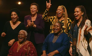 Six women singing and clapping on a stage, with a spotlight above.