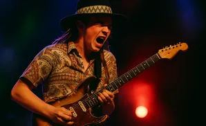 A guitarist in a hat and patterned shirt plays with passion under stage lights.