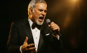 A man in a tuxedo sings into a microphone with his eyes closed and mouth open in a dramatic pose.