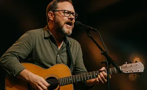 A man with glasses and a beard sings into a microphone while playing an acoustic guitar on stage.