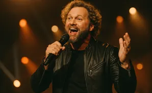 A man with curly hair and a beard, wearing a leather jacket, sings into a microphone with his hand raised.