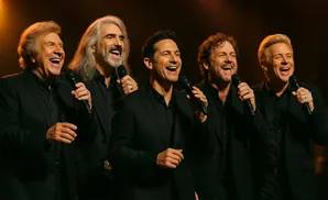 Five men in black suits singing into microphones on stage.