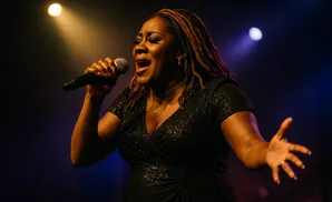 Singer with dreadlocks in a sparkly black dress sings into a microphone under stage lights.