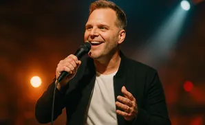 A man with short blonde hair smiles and holds a microphone, wearing a black jacket over a white t-shirt.