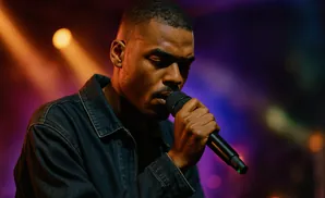 A man in a denim jacket sings into a microphone under purple and orange lights.