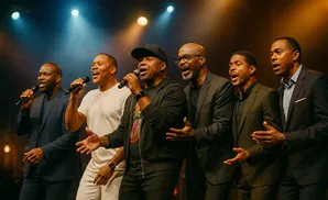 A group of six Black men singing on stage, lit by spotlights.