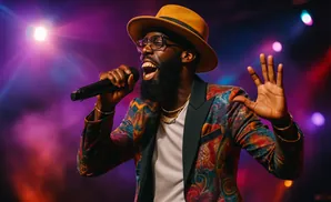 A Black man with a beard and glasses sings into a microphone, wearing a colorful patterned suit and a tan hat under stage lights.