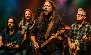 A band of five men performing on stage, with four singing and playing guitars and one in the background on drums.