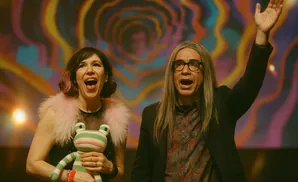 A woman holds a frog toy while a man with long hair and glasses raises his hand, both with mouths open in excitement.