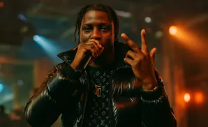 A Black man in a purple puffer jacket performs on stage with a microphone.