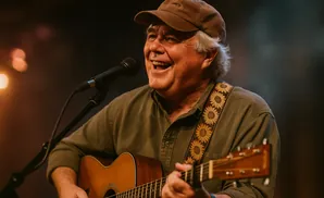 An older man with white hair wears a brown baseball cap and plays an acoustic guitar, singing into a microphone.
