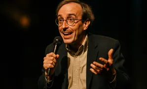 Man in glasses and suit speaks into a microphone, gesturing with his hands.