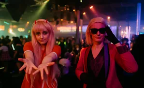 Two women pose for a photo at a party. The woman on the left has pink hair and devil horns, while the woman on the right wears sunglasses and a red blazer.