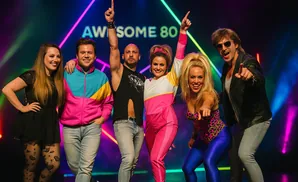 Group of five people in 80s attire posing with bright neon lights in the background.
