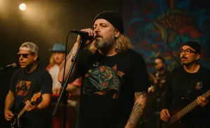 A man with a beard and blonde hair sings into a microphone at a concert, wearing a t-shirt with a fish design.