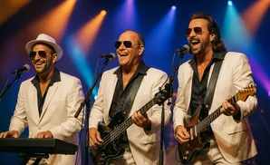 A band in white suits sings and plays instruments on stage under blue and orange spotlights.