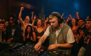 A DJ plays music for a lively crowd with raised hands and smiles.